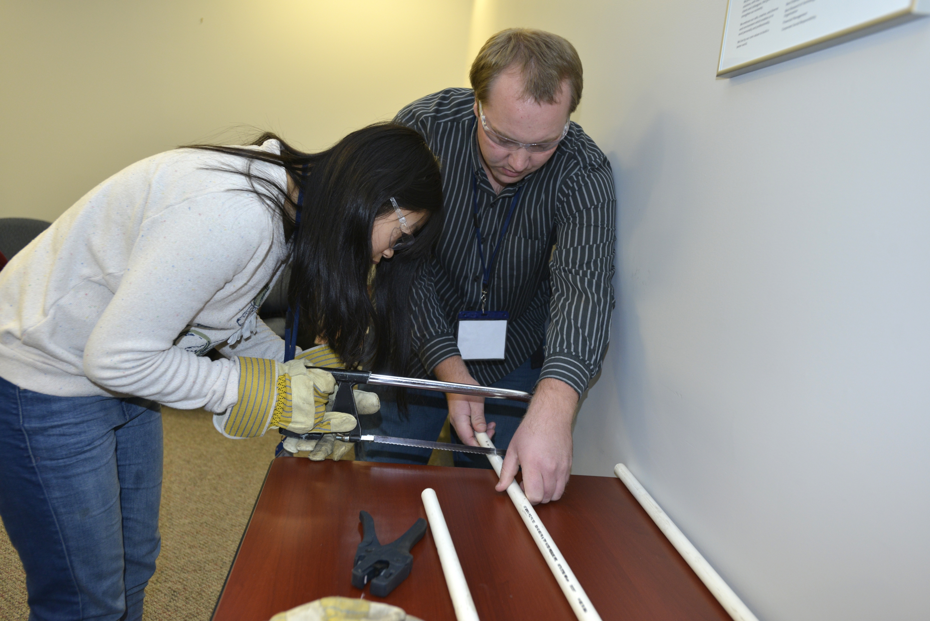 student wears safety gloves and glasses while sawing a PVC pipe held by instructor