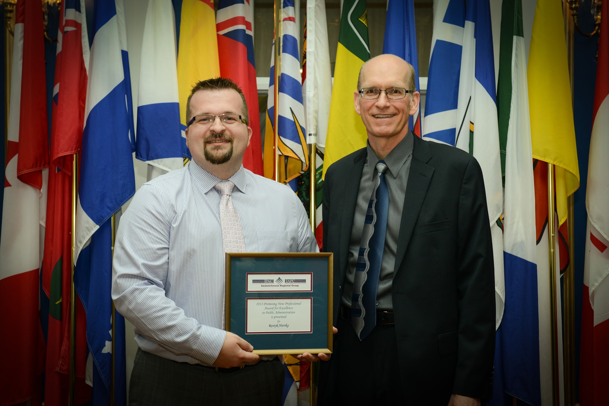 two men stand in front of flags, one holding a plaque