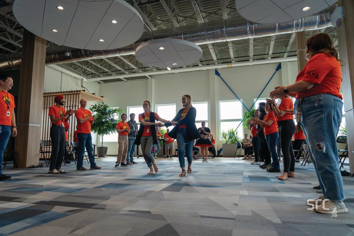 A group of people wearing orange shirts gathered in a circle inside a modern, well-lit indoor space with high ceilings. Two women hold hands and dance in the center of the room while others around them clap and smile. The event appears to be an Orange Shirt Day or Every Child Matters gathering