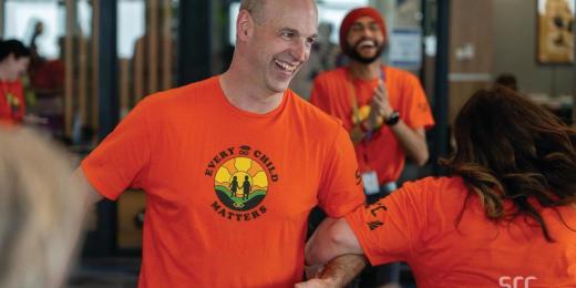 A smiling man wearing an orange 'Every Child Matters' t-shirt featuring a sunrise and silhouettes of two children holding hands. He is engaged in conversation with others, while a smiling man wearing a red turban and matching orange shirt claps in the background. The setting appears to be a bright indoor office or community space.
