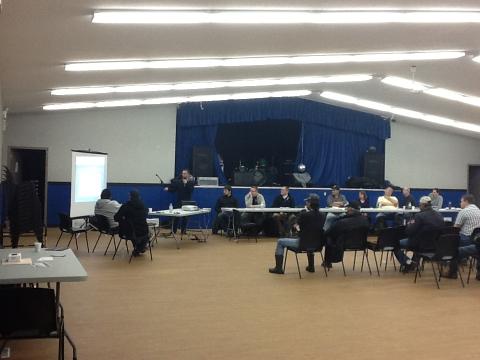 People sitting in a u-shape around a projector screen in a large hall