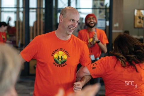 A smiling man wearing an orange 'Every Child Matters' t-shirt featuring a sunrise and silhouettes of two children holding hands. He is engaged in conversation with others, while a smiling man wearing a red turban and matching orange shirt claps in the background. The setting appears to be a bright indoor office or community space.