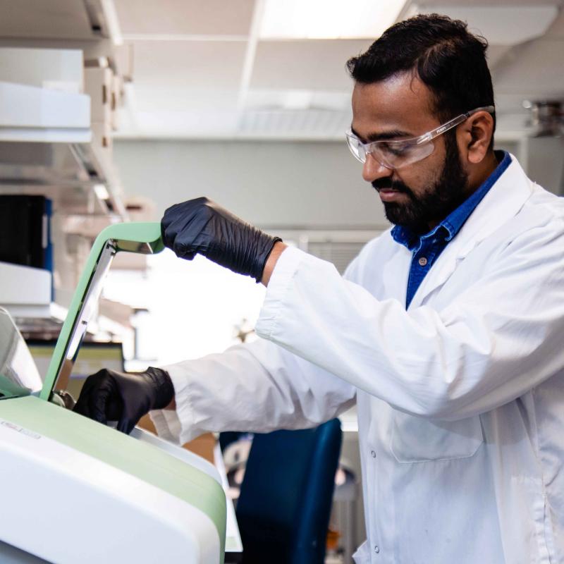 A scientist wearing a white lab coat, safety glasses, and black nitrile gloves operates a piece of laboratory equipment with an open green lid in a bright, modern lab.