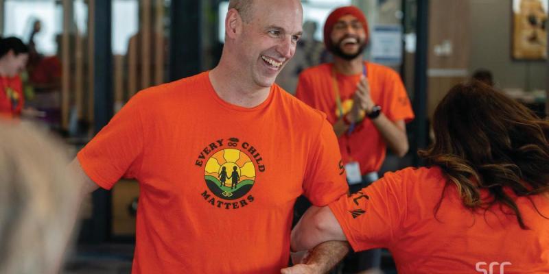 A smiling man wearing an orange 'Every Child Matters' t-shirt featuring a sunrise and silhouettes of two children holding hands. He is engaged in conversation with others, while a smiling man wearing a red turban and matching orange shirt claps in the background. The setting appears to be a bright indoor office or community space.